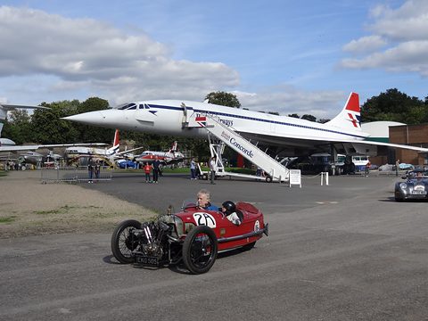 A small red three-wheeler on tarmac in the foreground, a Concorde airliner parked behind it under a blue sky. Opens a larger view