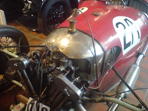 Close view of a polished tank and an air-cooled V-twin with pipes on a red racing three-wheeler numbered 28. Opens a larger view