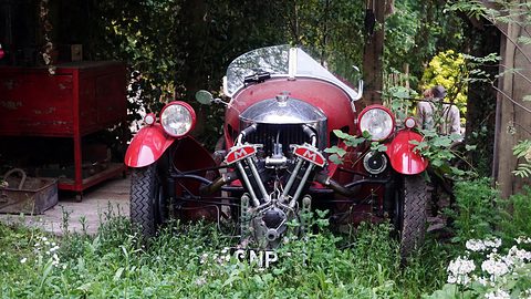 A red three-wheeler with an exposed V-twin and two headlamps standing among flowers and greenery in a garden set. Opens a larger view
