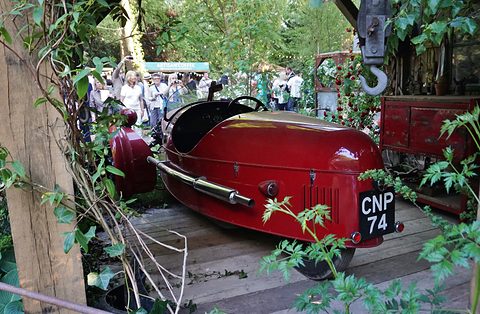 The rounded red tail of a three-wheeler with its number plate, in a garden of plants, visitors behind. Opens a larger view