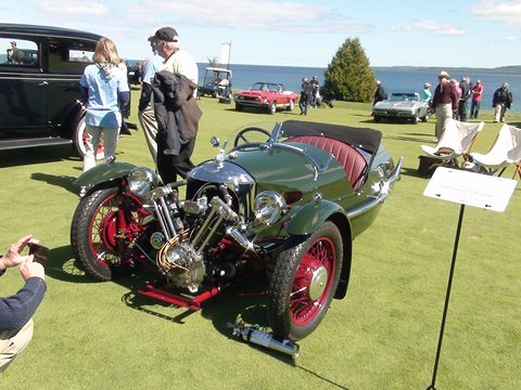 A dark green three-wheeler with red wheels and exposed V-twin on a lawn by a lake, visitors around it. Opens a larger view