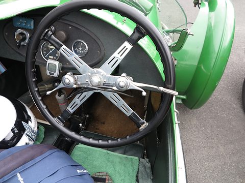 Looking down at a black four-spoke steering wheel, dashboard dials and a green body, a helmet and bag on the seat. Opens a larger view