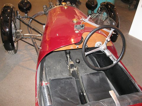 Looking down into a red three-wheeler: wooden dashboard, steering wheel, gear lever and two black seats. Opens a larger view