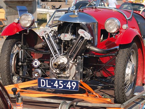 Front view of a red three-wheeler with a polished cowl, V-twin and Dutch plate, orange straps holding it on a trailer. Opens a larger view