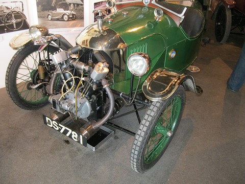 A green three-wheeler with brass headlamps, exposed V-twin and green wire wheels on a grey floor. Opens a larger view
