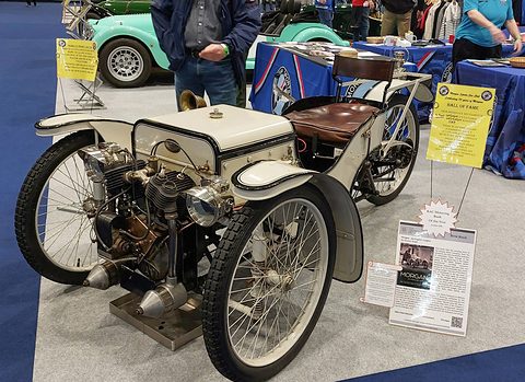 A white early three-wheeler with exposed V-twin, box bonnet and brown leather seat on grey carpet, a placard beside it. Opens a larger view
