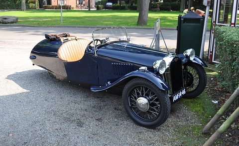 A dark blue three-wheeler with a radiator grille in front, swept wings, wire wheels and tan seats, on gravel. Opens a larger view