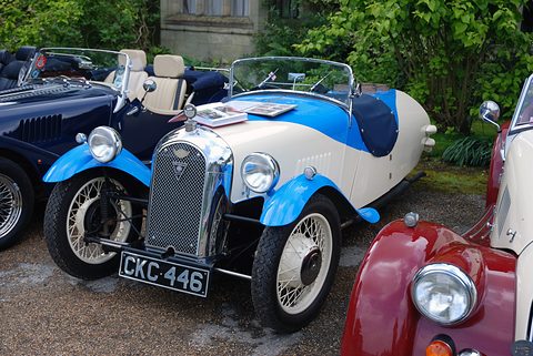A cream and bright blue three-wheeler with a mesh grille, two headlamps and black seats among other Morgans. Opens a larger view