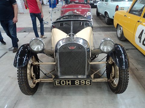 Front view of a cream and black three-wheeler with a mesh radiator grille, two headlamps and a number plate. Opens a larger view