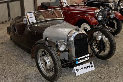 A dark brown four-seat three-wheeler with a chrome grille and wire wheels, a Bonhams lot card and sign, a red car behind. Opens a larger view