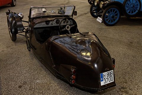 Rear quarter view of a dark brown three-wheeler with a rounded tail and a Spanish number plate. Opens a larger view