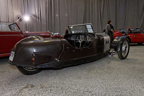 Side view of a dark brown three-wheeler with a rounded tail, before a grey curtain and other sale cars. Opens a larger view