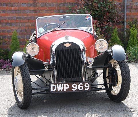Front view of a red three-wheeler with an upright chrome grille, two headlamps and a black number plate, brick wall behind. Opens a larger view