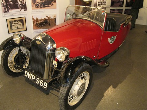 A red four-seat three-wheeler with a chrome grille, black wings and a folded hood before framed photographs. Opens a larger view