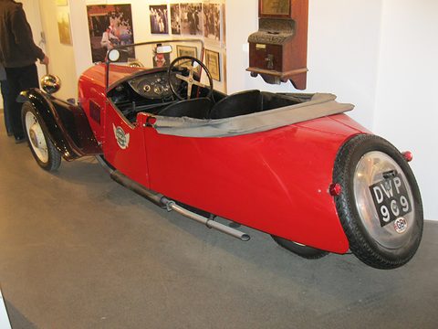 Rear quarter view of a red three-wheeler with a spare wire wheel on its tail and a folded hood. Opens a larger view
