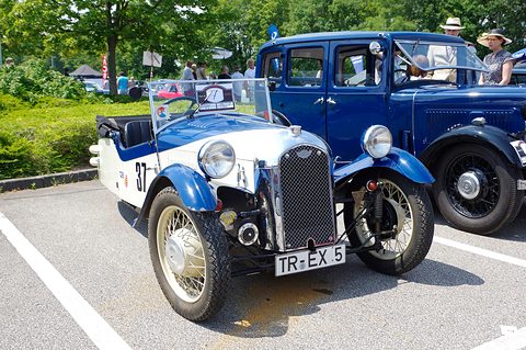 A cream and blue three-wheeler with a chrome grille and a rally start card on its windscreen, a dark blue saloon behind. Opens a larger view