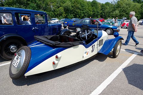 A cream and blue three-wheeler with a spare wheel on its tail, parked between white lines with other old cars around. Opens a larger view
