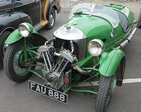 A green three-wheeler with exposed V-twin, two headlamps and green wire wheels parked on tarmac. Opens a larger view