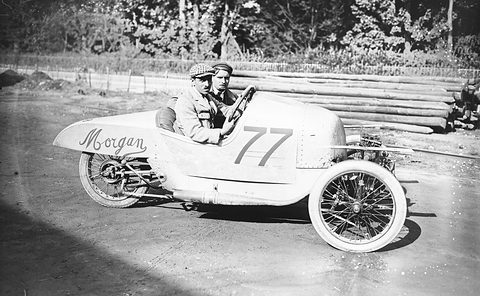 Two men in caps in a pale pointed-tail three-wheeler numbered 77, timber stacked behind on a dirt yard.