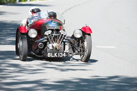 A red three-wheeler with exposed V-twin and two helmeted occupants on a sunlit mountain road. Opens a larger view