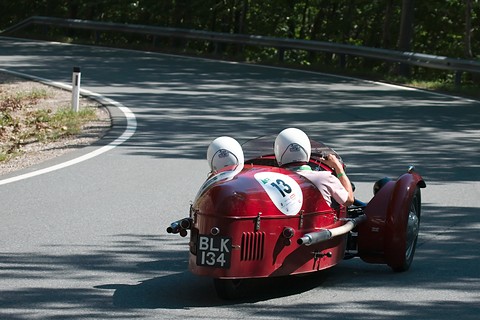 Rear view of a red three-wheeler with two helmeted occupants rounding a bend on a mountain road. Opens a larger view
