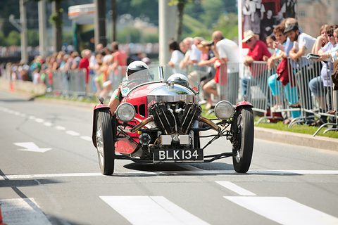 A red three-wheeler with exposed V-twin driving on a city street past spectators behind metal barriers. Opens a larger view