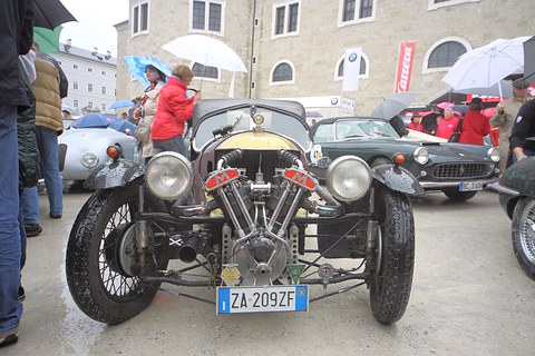 Front view of a dark three-wheeler with exposed V-twin and two headlamps on a wet square, people with umbrellas behind. Opens a larger view