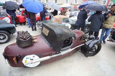 Looking down on a maroon three-wheeler with a leather bag strapped to its tail, people with umbrellas around it. Opens a larger view