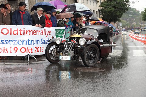 A dark three-wheeler on a wet street before spectators with umbrellas behind a rally banner. Opens a larger view