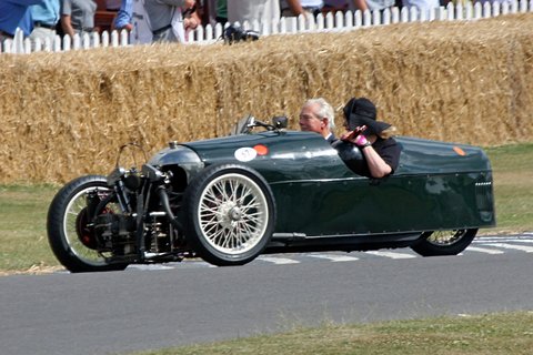 A dark green beetle-backed three-wheeler with two people aboard drives past straw bales and spectators. Opens a larger view