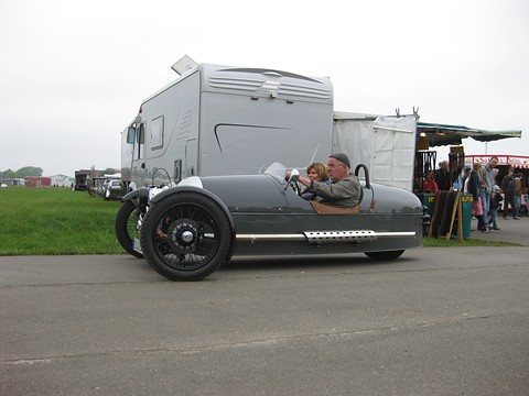 Side view of a grey three-wheeler with two people in it on tarmac, a motorhome and market stalls behind. Opens a larger view