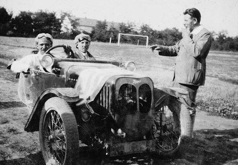 Two smiling men in leather caps seated in a small three-wheeler with wire wheels, a man in a jacket pointing beside it. Opens a larger view