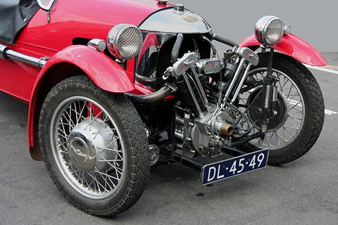 Low front view of a red three-wheeler showing the V-twin, two headlamps, wire wheels and a Dutch number plate. Opens a larger view