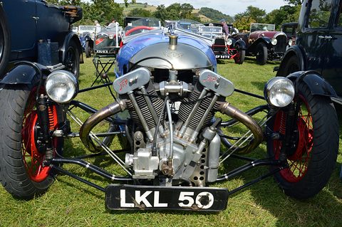 Front view of a blue three-wheeler with a large JAP V-twin, two headlamps and red wheels on grass at a show. Opens a larger view