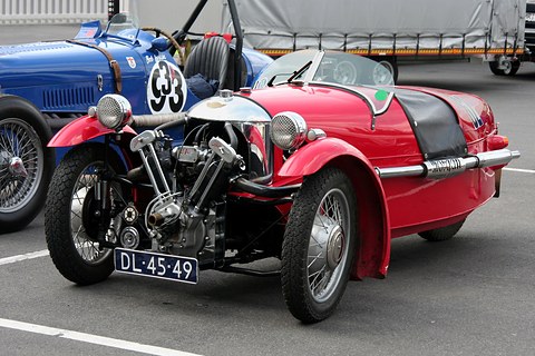 A red three-wheeler with a polished cowl, exposed V-twin and black tonneau on tarmac, a blue racing car numbered 33 behind. Opens a larger view