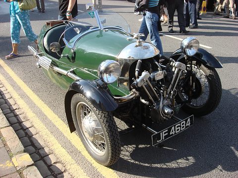 Looking down on a green three-wheeler with a polished cowl and exposed V-twin by a kerb with double yellow lines. Opens a larger view