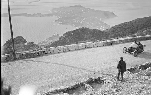 A small three-wheeler with two crew on a mountain road edged by a stone wall, a man in a hat watching, the coast far below. Opens a larger view