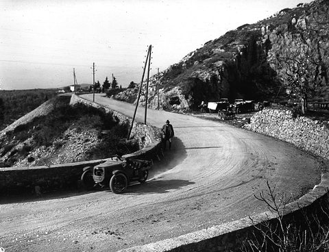 A small open three-wheeler rounding a walled hairpin bend on a dusty mountain road, a man standing at the verge above. Opens a larger view