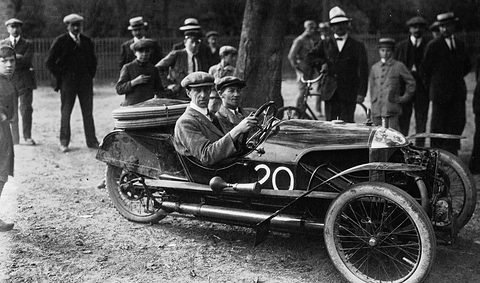 Side view of a long dark three-wheeler numbered 20 with exhaust pipes along its flank, a crowd of men and boys behind. Opens a larger view