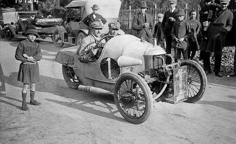 A driver in a pale three-wheeler numbered 1 with exposed V-twin, a girl in a dark dress and men in caps around it. Opens a larger view