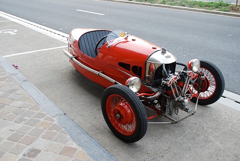 Looking down on a red three-wheeler with exposed V-twin and red wire wheels parked at a kerb. Opens a larger view