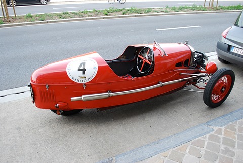 Side view of a red three-wheeler with a racing number 4, a long exhaust pipe and a black leather seat, by a road. Opens a larger view