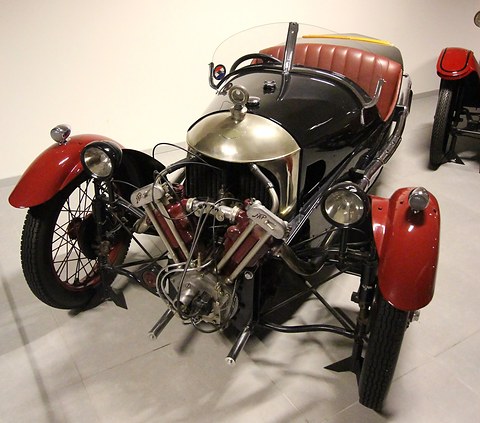 Front view of a black three-wheeler with red wings, a polished cowl and an exposed V-twin with red cylinder heads. Opens a larger view