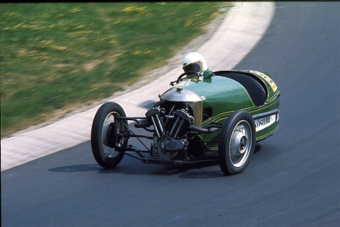A green three-wheeler with an exposed V-twin and a helmeted driver cornering on a track beside a grass verge. Opens a larger view