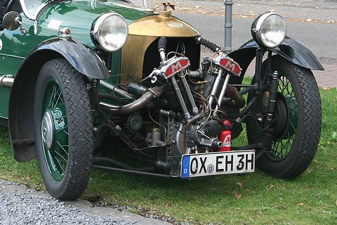 A green three-wheeler with brass cowl, two headlamps and a Matchless V-twin, parked on grass by a road. Opens a larger view