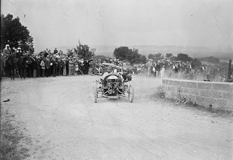 A three-wheeler with an exposed V-twin heads up a dusty bend, a woman passenger leaning out, spectators on horseback and foot behind. Opens a larger view