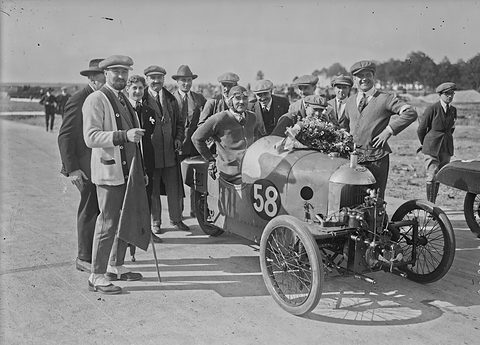 A man sniffing a bouquet in a small three-wheeler numbered 58, surrounded by men in caps and coats. Opens a larger view