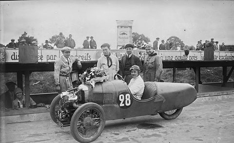 A moustached man in a leather helmet and goggles in a pointed-tail three-wheeler numbered 28 with flowers on its bonnet, men behind a barrier.