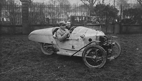 A pale pointed-tail three-wheeler with two men aboard on grass before iron railings and bare trees. Opens a larger view