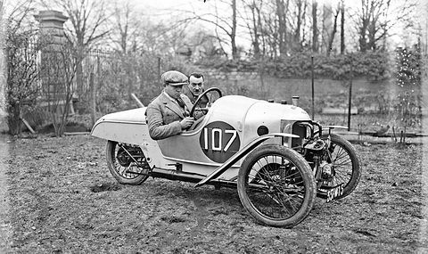 Two men in a pale three-wheeler numbered 107 on rough ground before bare trees and a gate pier. Opens a larger view